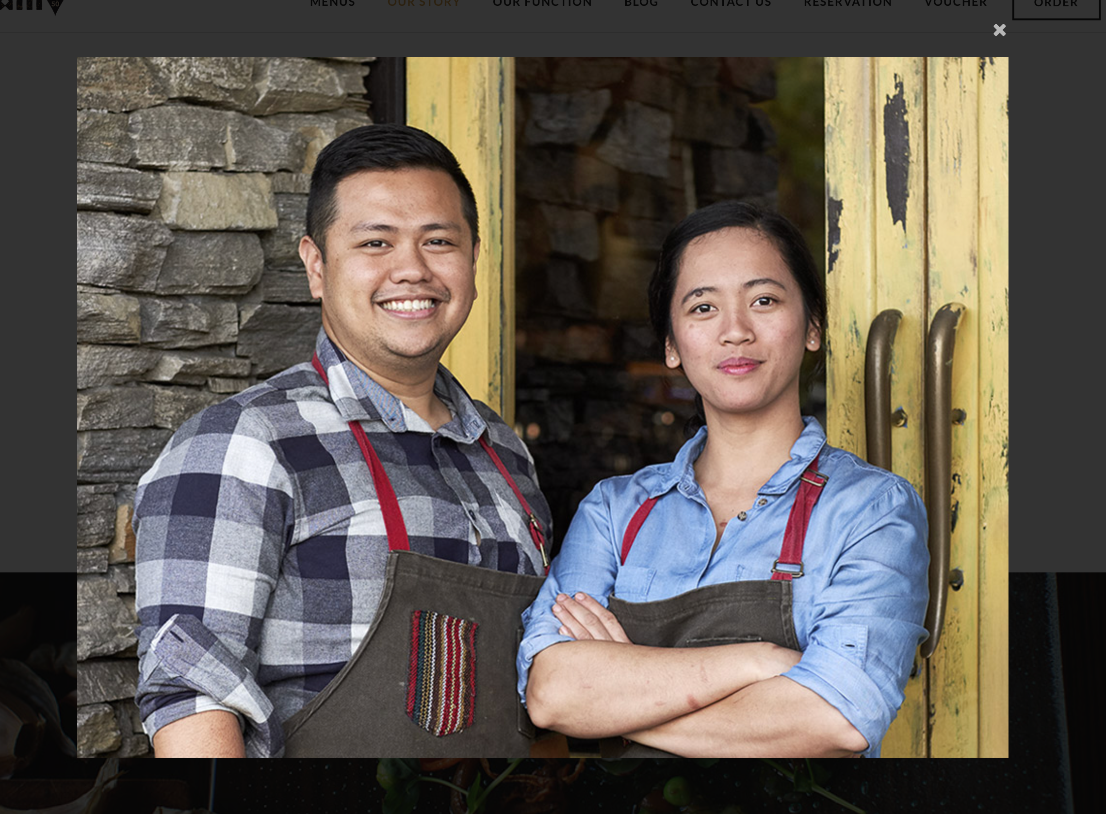 Two chefs of Filipino descent wearing grey aprons with red straps standing in front of a closed restaurant