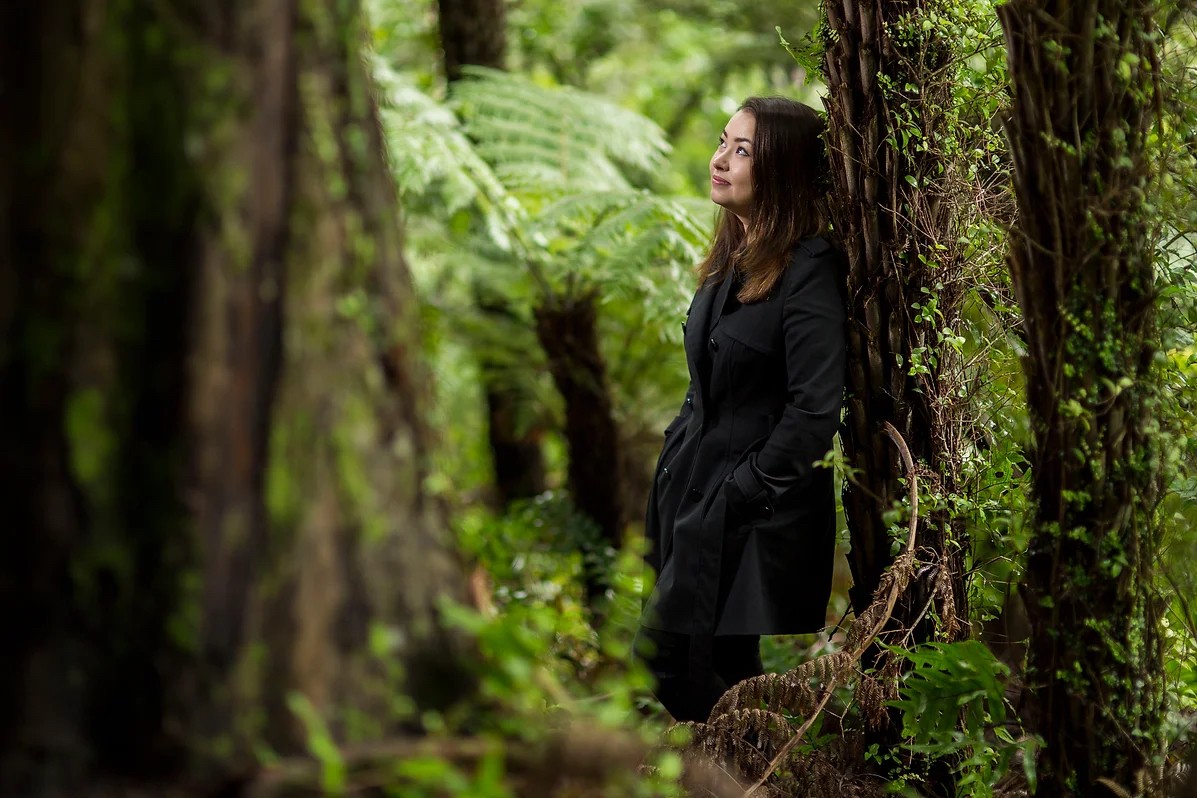 A woman with dark shoulder-length hair leans against a ponga trunk and gazes up at the bush