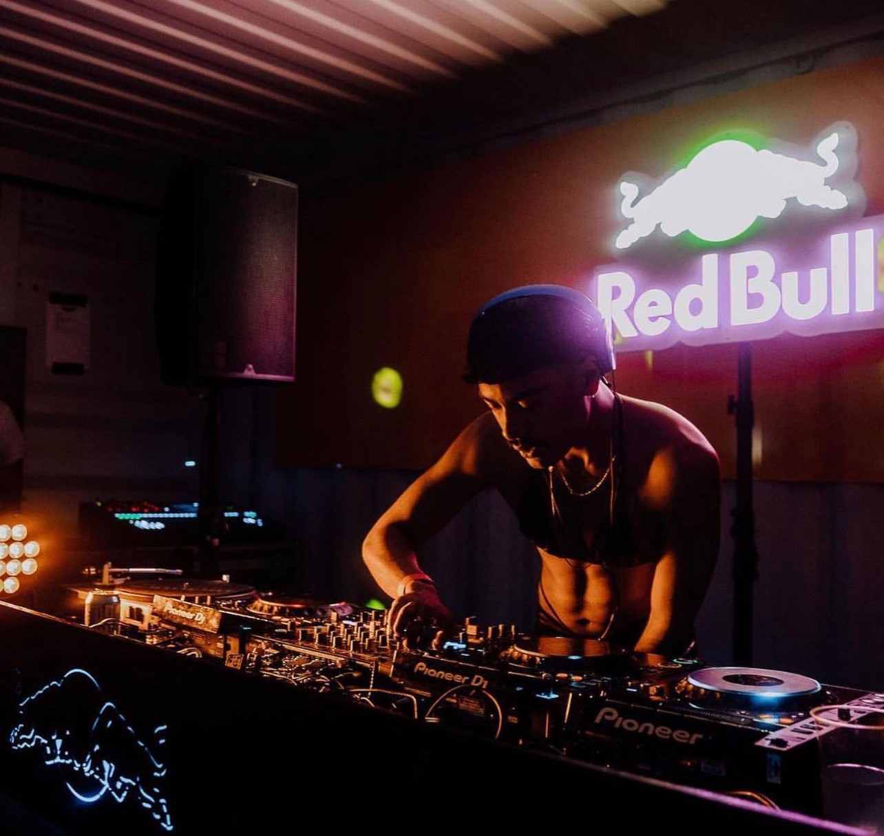 A dimly lit DJ booth with a huge Red Bull sign with a dj in a halter top performing