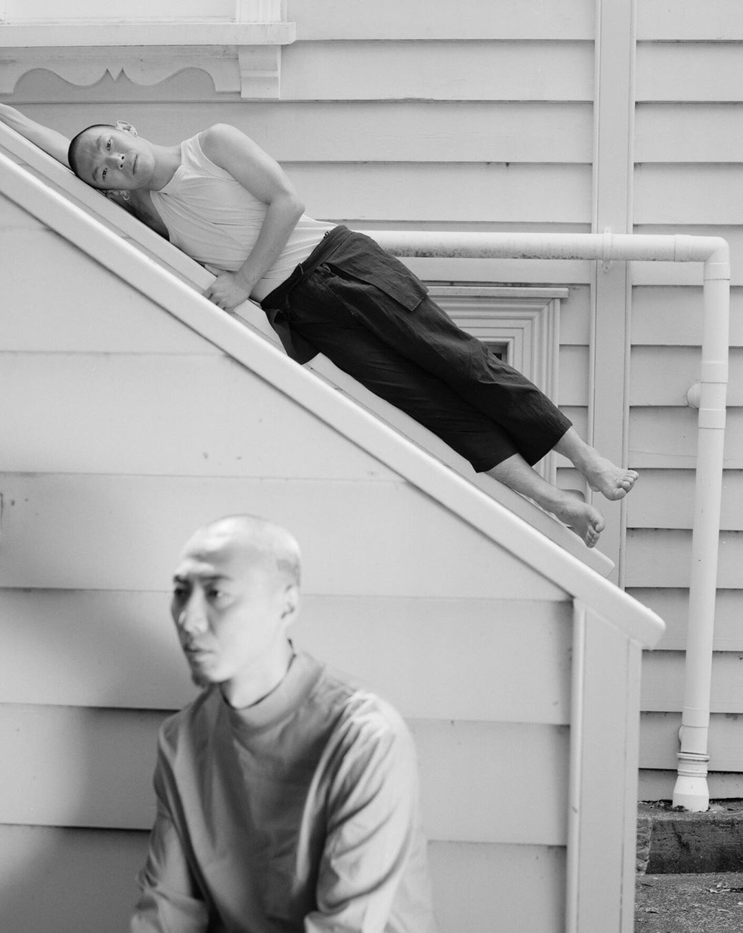 Black and white photograph of two dancers, one sitting in the foreground, the other lying on an angled roof.