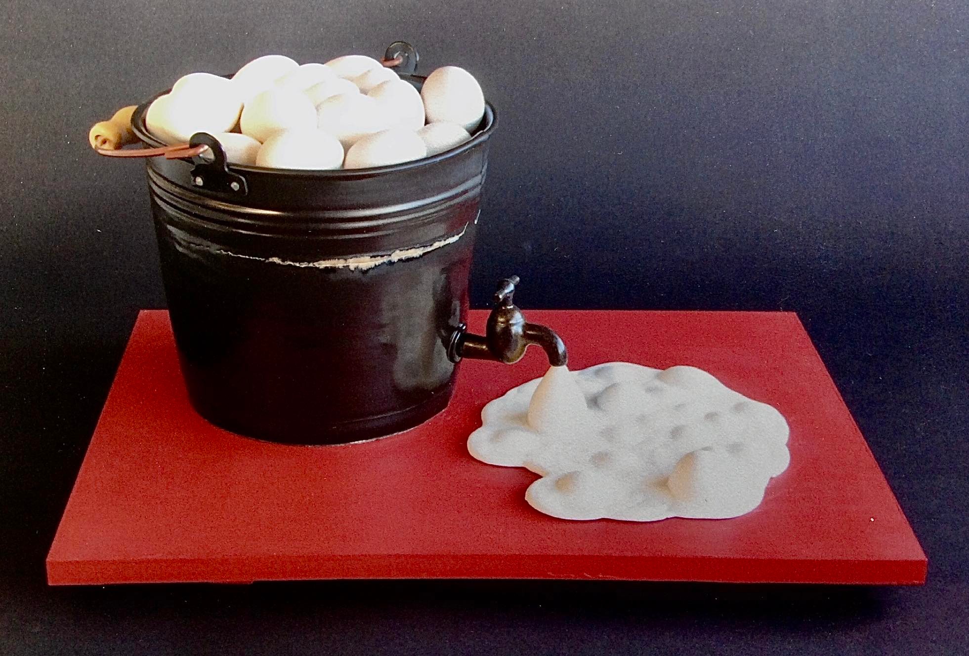 A sculpture of eggs in black bucket with a spout that has milk coming out of it. 