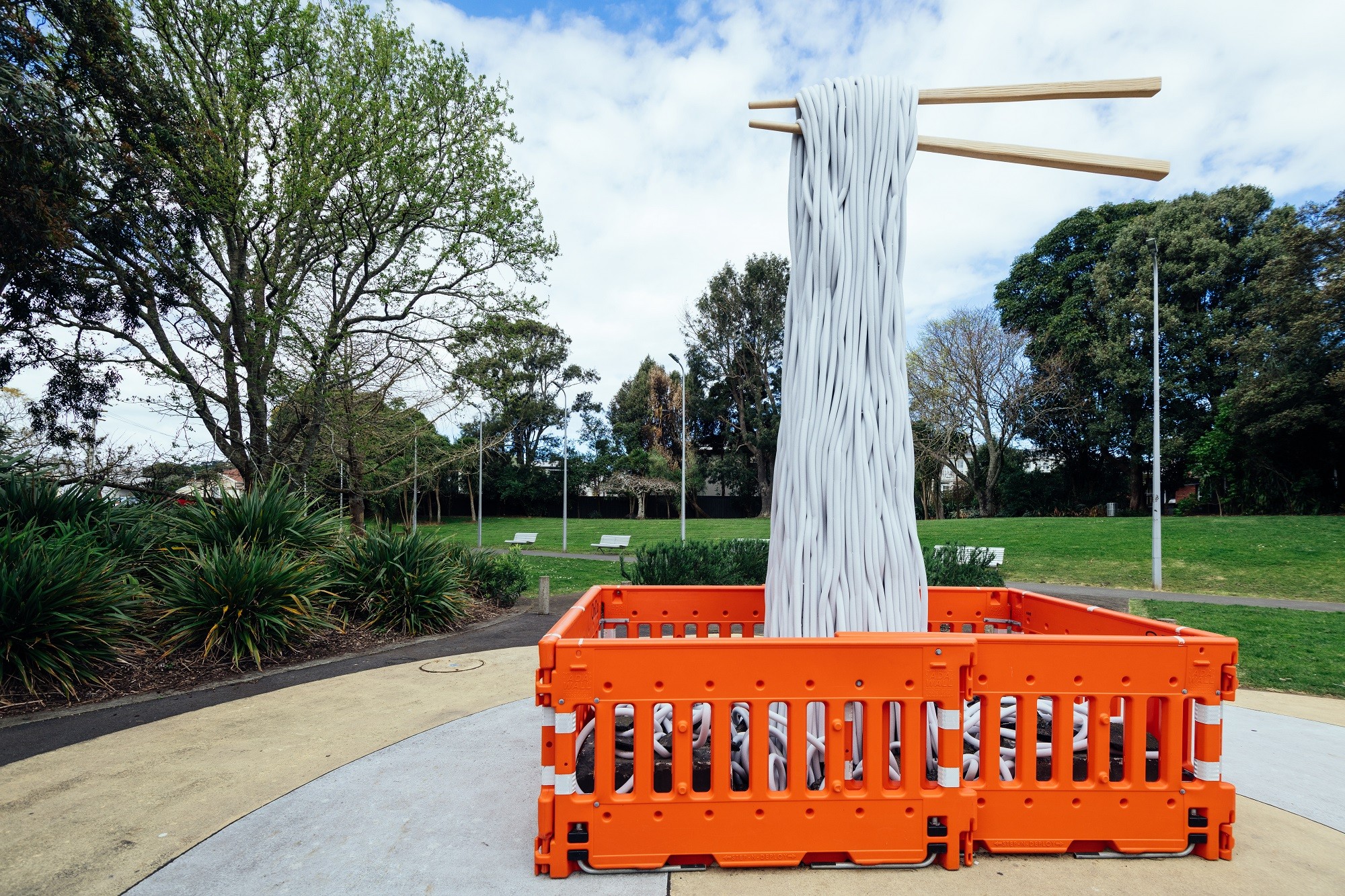 Orange roadwork barricades surrounding a tower of noodle-like objects being lifted with a giant pair of chopsticks by an invisible hand. In the background is a pathway with bench seats, lampposts, and a grassy area with trees behind it. 