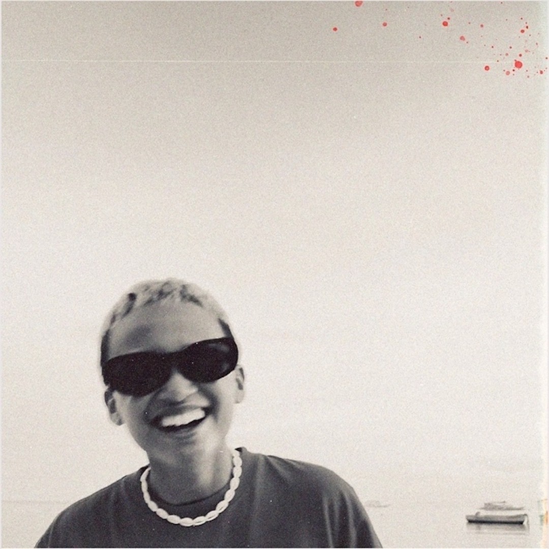 Sepia-toned photo of a smiling Filipino person with short hair wearing dark sunnies and a shell necklace