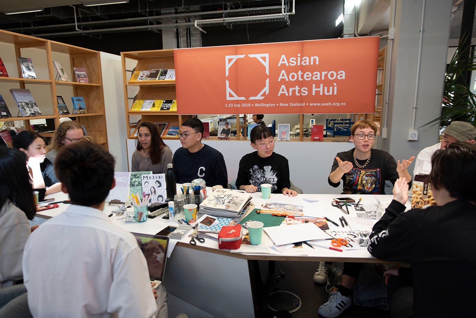 A group of people sit around a table with zine materials in front of a zine display.