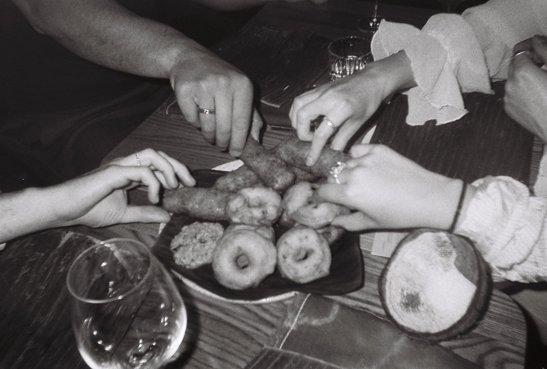 Grayscale photo of four hands reaching for a shared plate of food on a table with drinking glasses