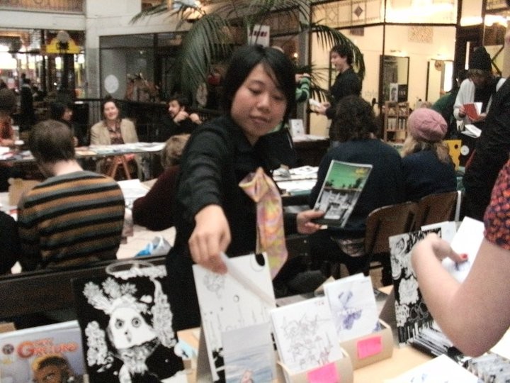 A person manning a zine table and showing zines to a customer. 