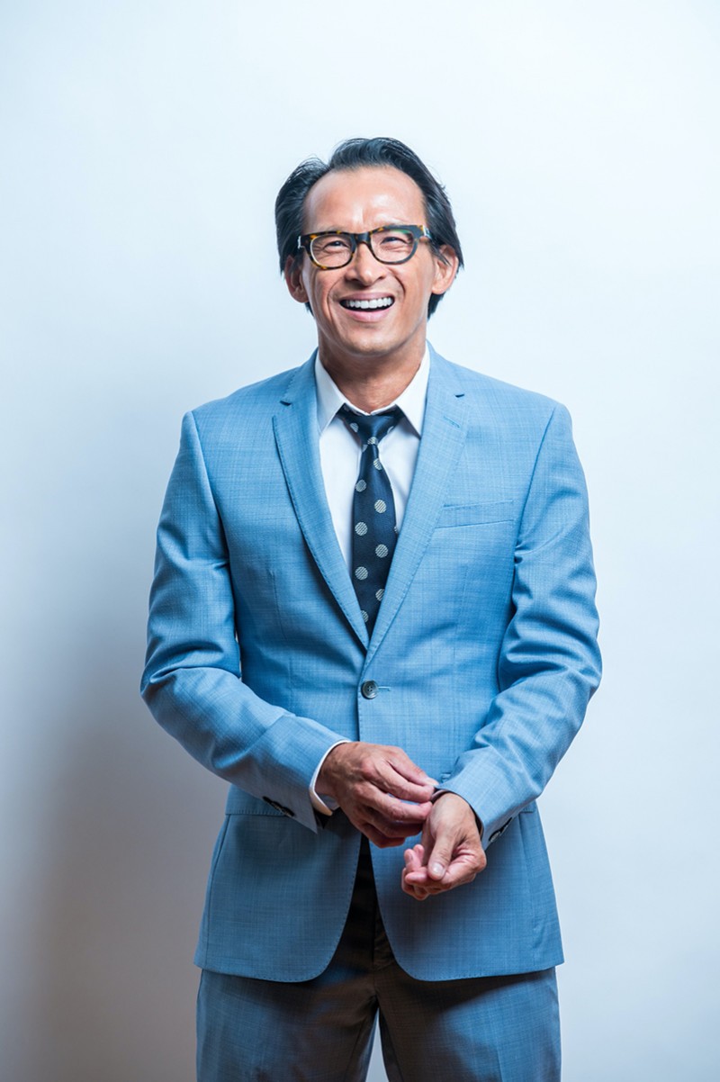 A middle-aged man in a powder blue suit and glasses laughing at the camera as he fixes one of his sleeves