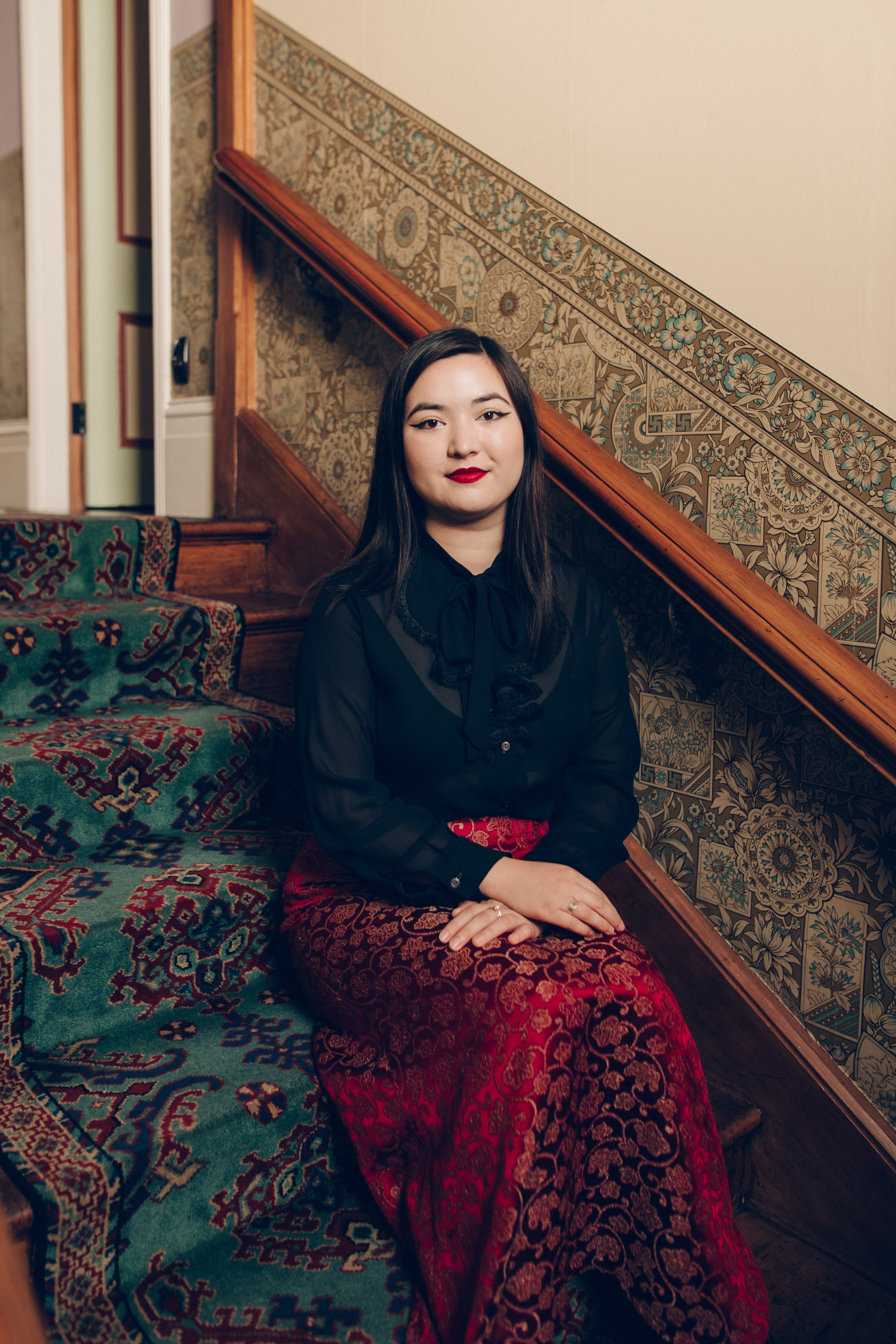 A woman with long black hair wearing a black lace sheer shirt and a red patterned maxi skirt sitting on an emerald carpeted staircase