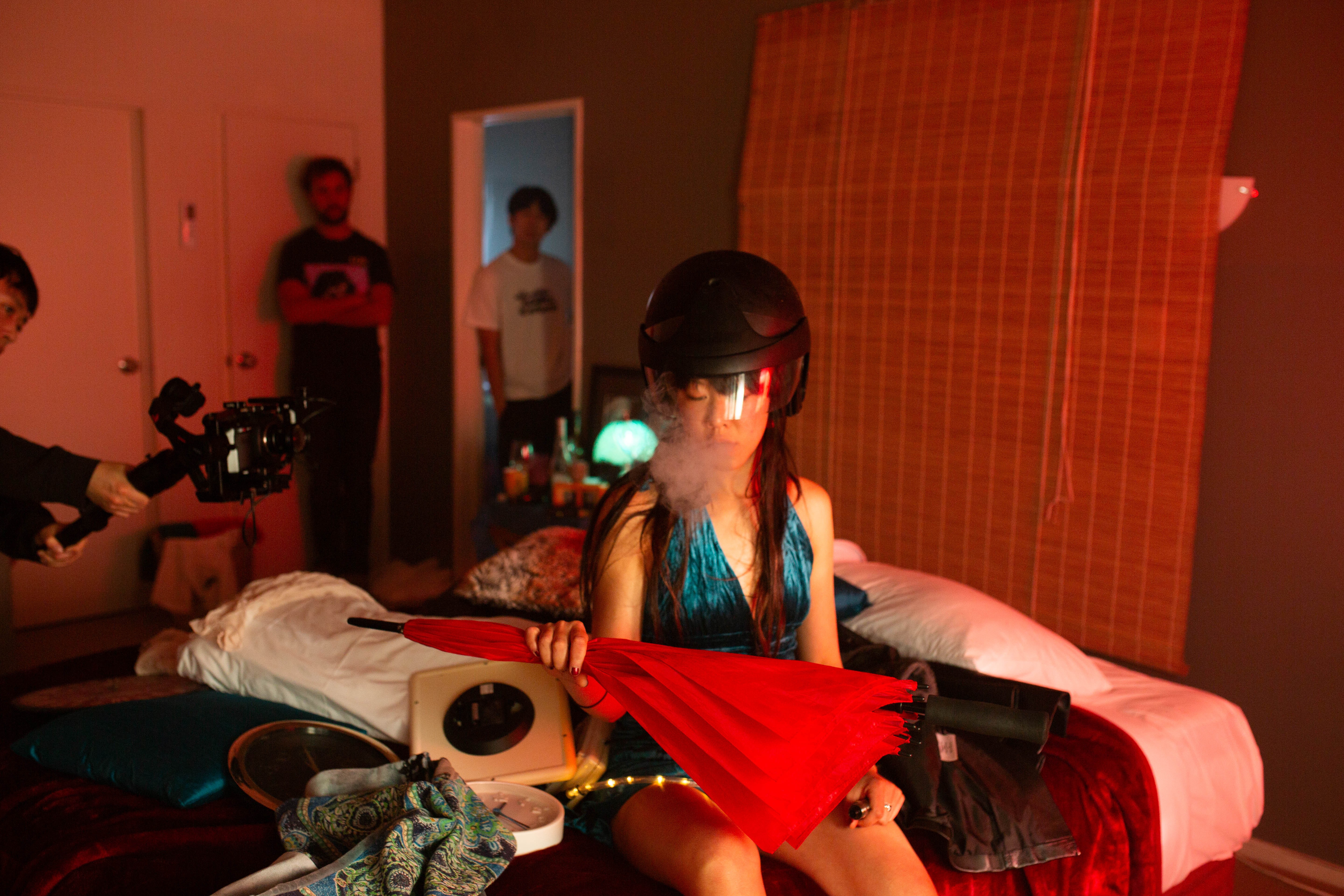 A woman sits on a red-lit bed holding an umbrella and wearing a motorcycle helmet surrounded by a film crew