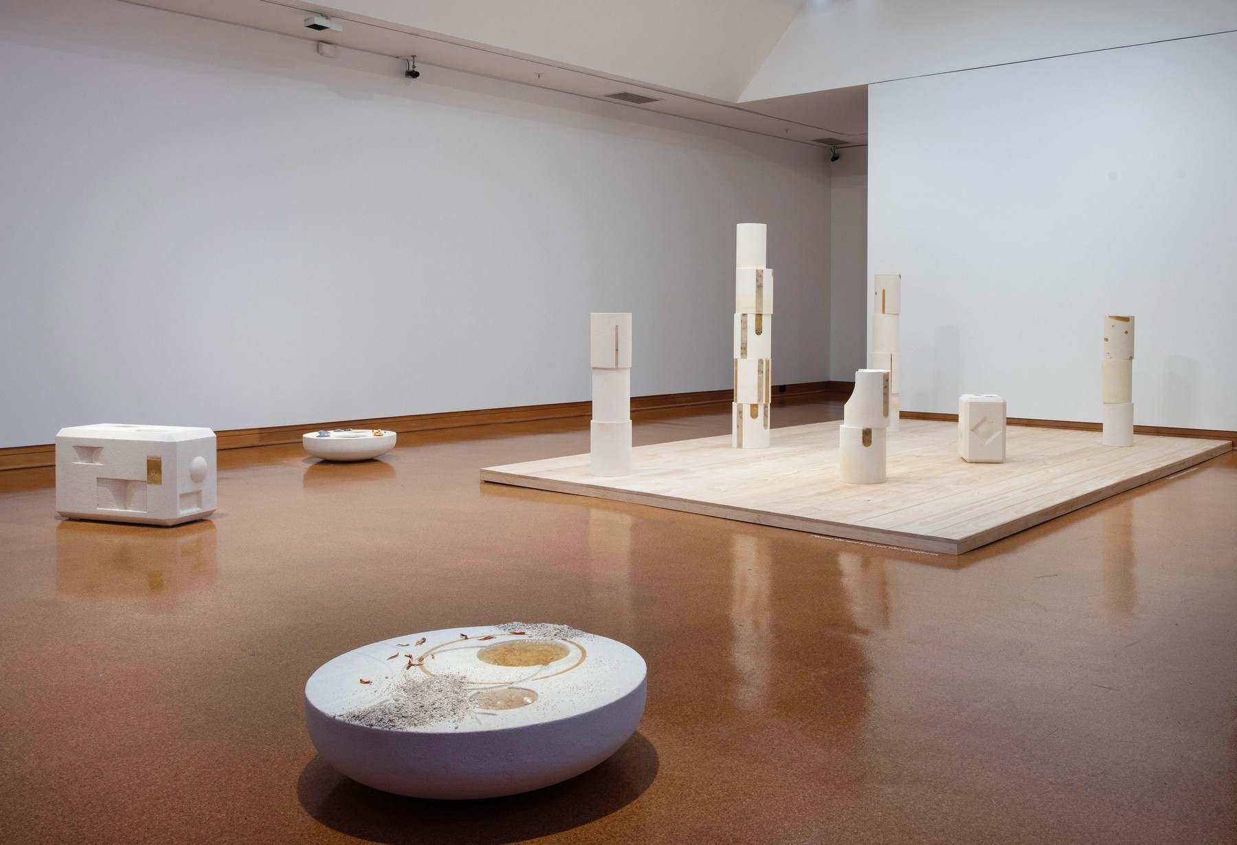 A series of white cast objects across the floor — two large circular plinths with, and five pillars of different heights on a plain MDF floor objects inlaid, a rectangular object with indents evoking a weight