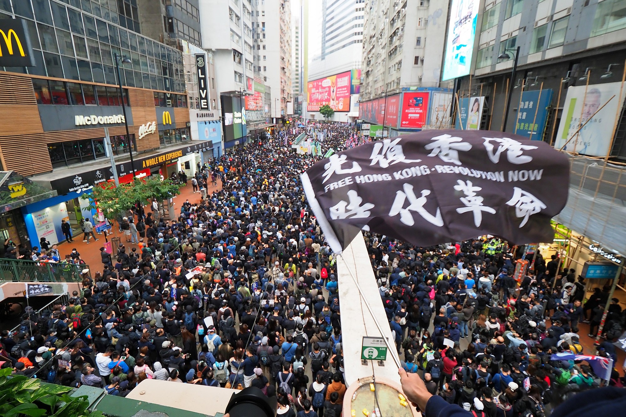 A flag with the Free Hong Kong slogan in English and Chinese flies high above a crowd of protestors on a street.