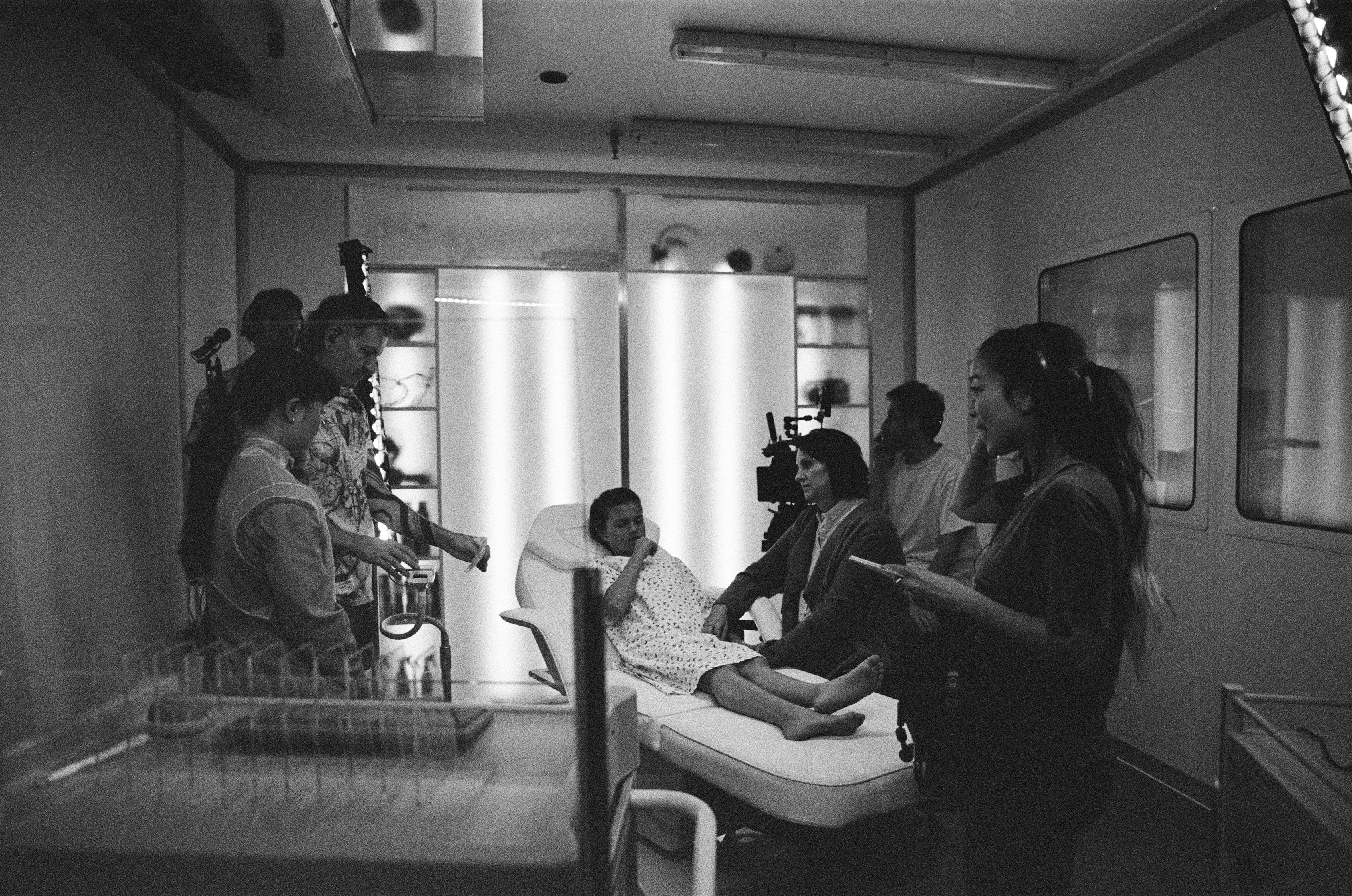 Black and white behind-the-scenes image of a film set, showing a woman in a hospital gown lying on a medical bed surrounded by crew members, including a director giving notes, a camera operator, and sound and lighting technicians.