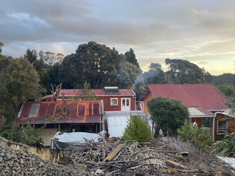 Orange and red corrugated iron roofs and a chimney emitting smoke with trees and a cloudy gold sky in the background and a pile of dry branches in the foreground