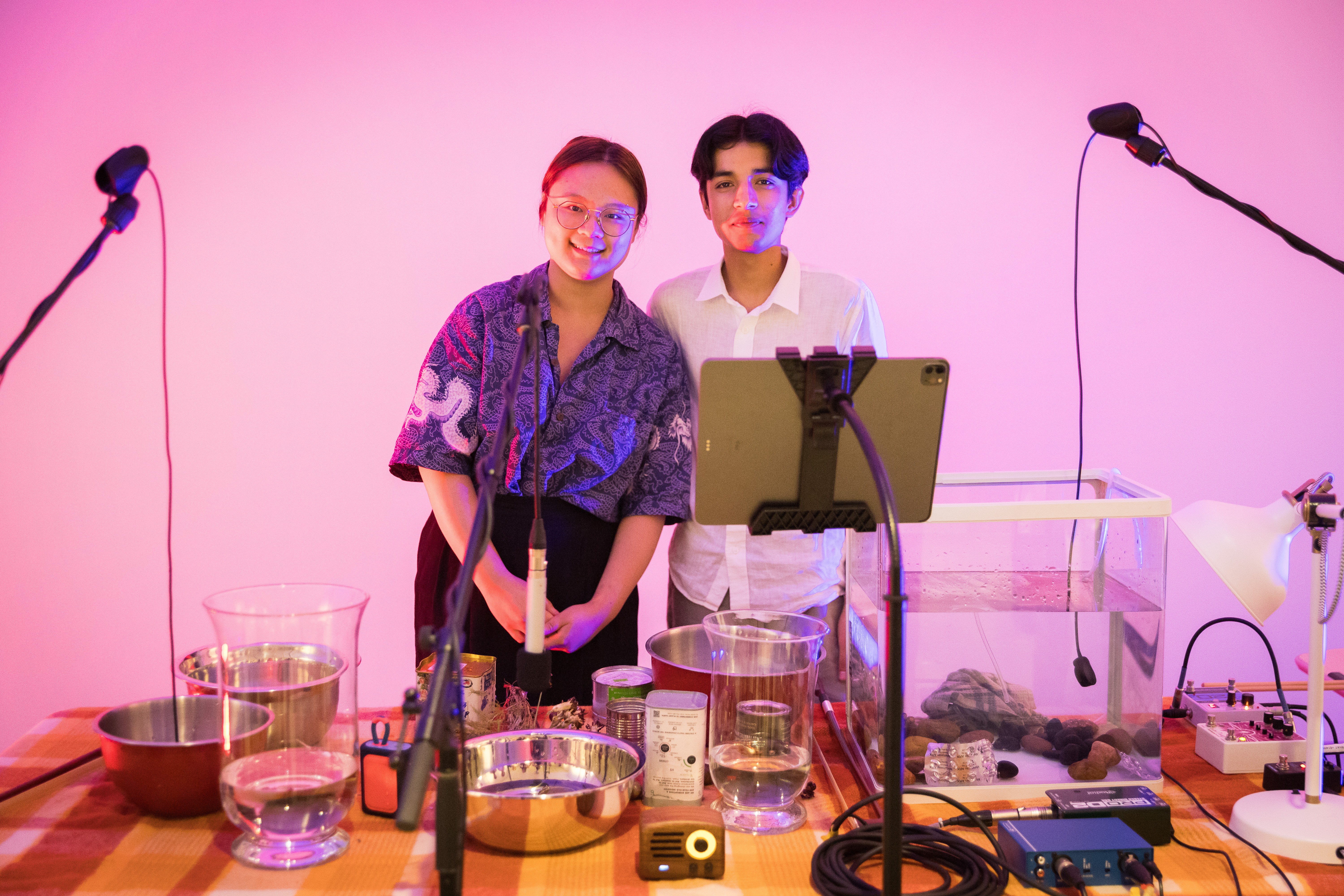 A Chinese person wearing a blue top smiles next to a South Asian man wearing a white shirt behind a table full of instruments.