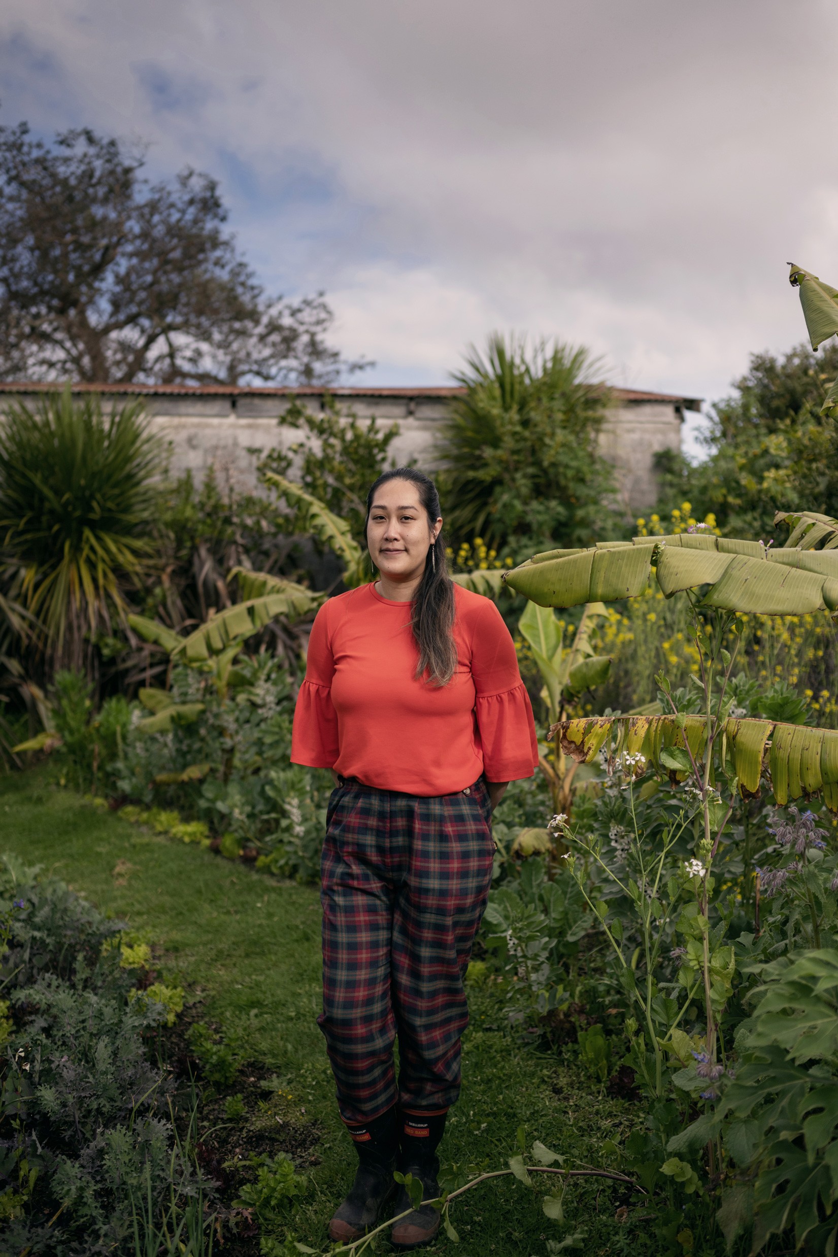 Person of Asian descent wearing a bright orange top standing in front of a lush garden