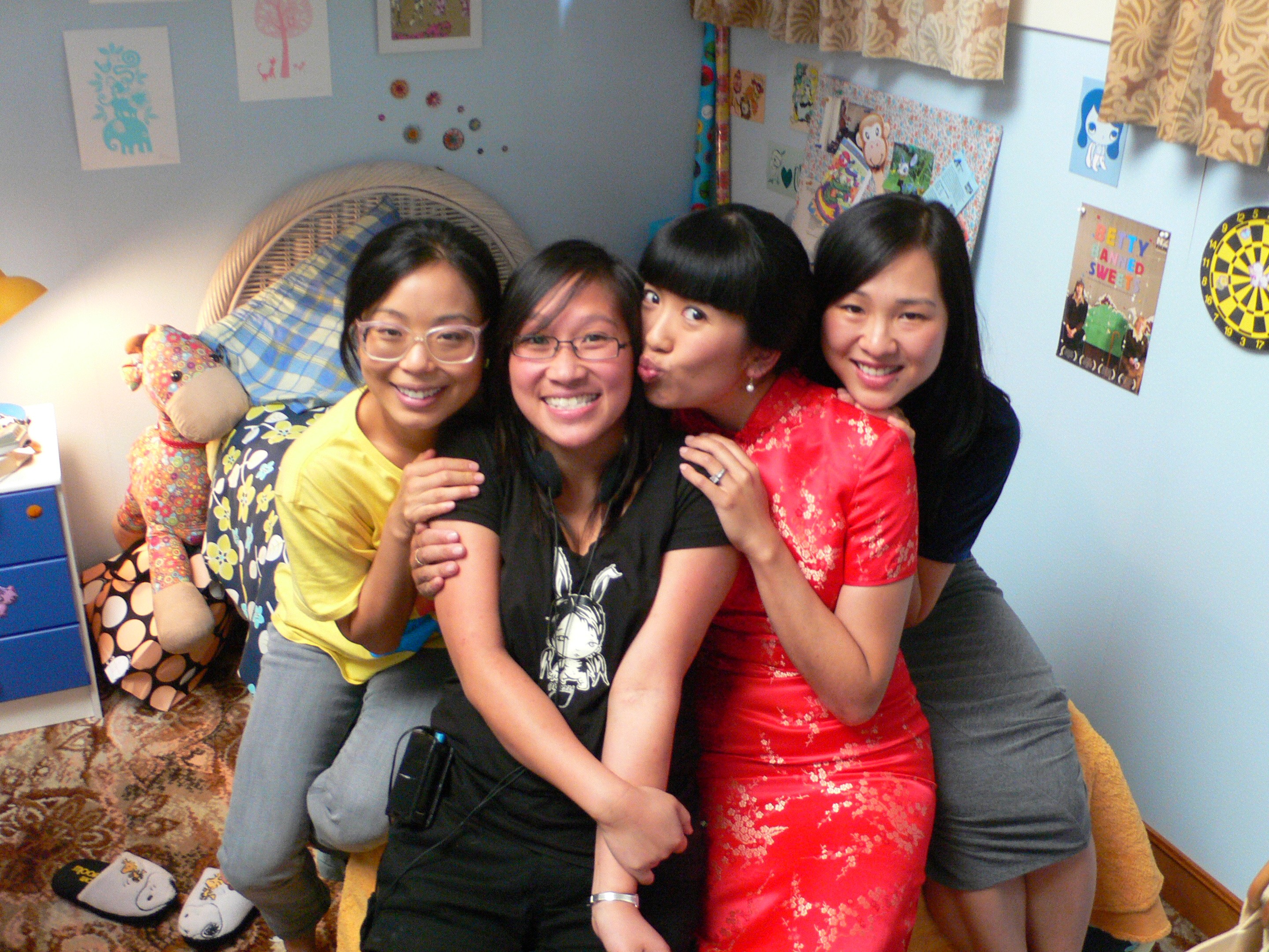 Four East Asian women perched on the edge of a bed in a teenager's bedroom smiling at the camera