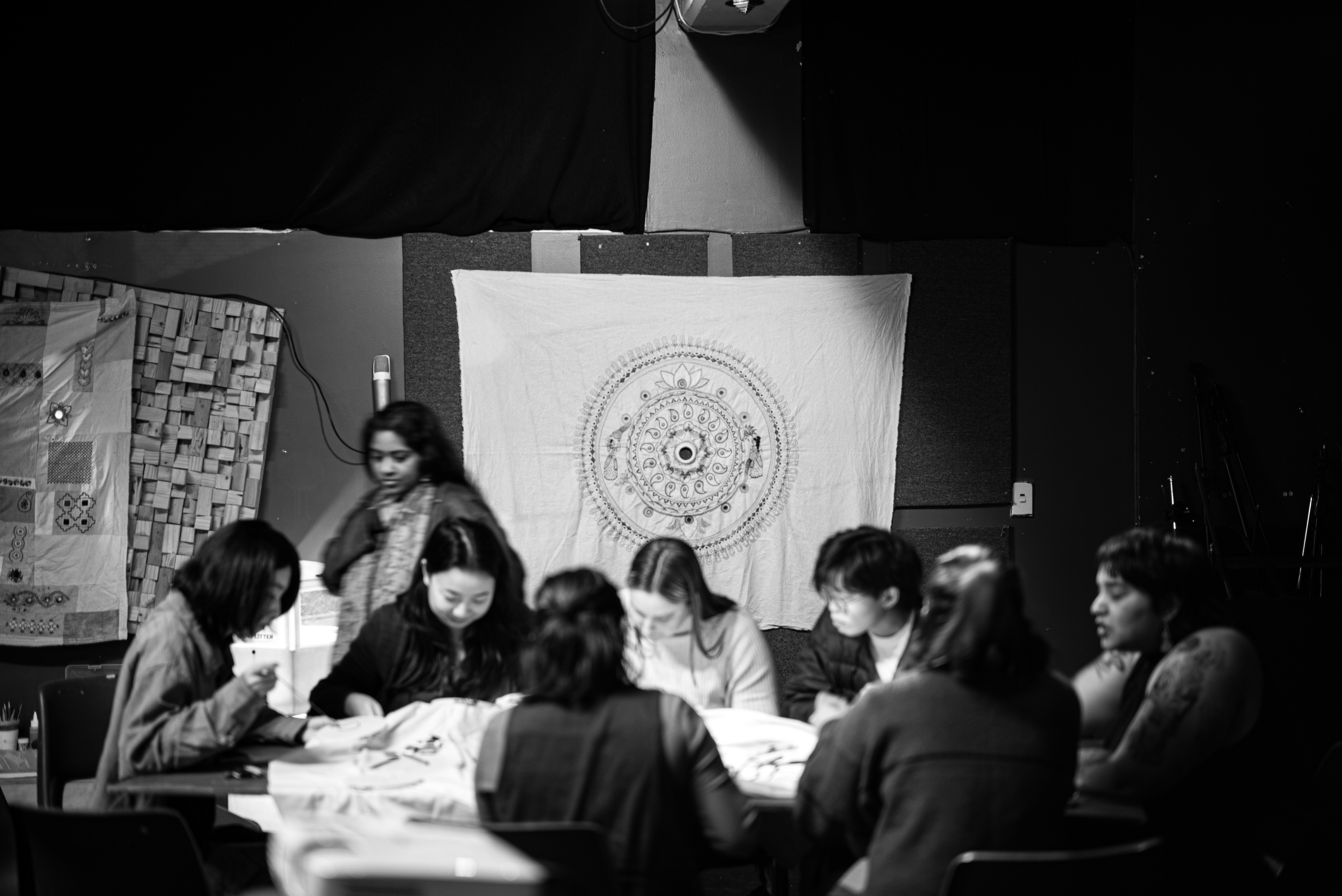 People sitting around a table with embroidered artwork hanging up behind them. 