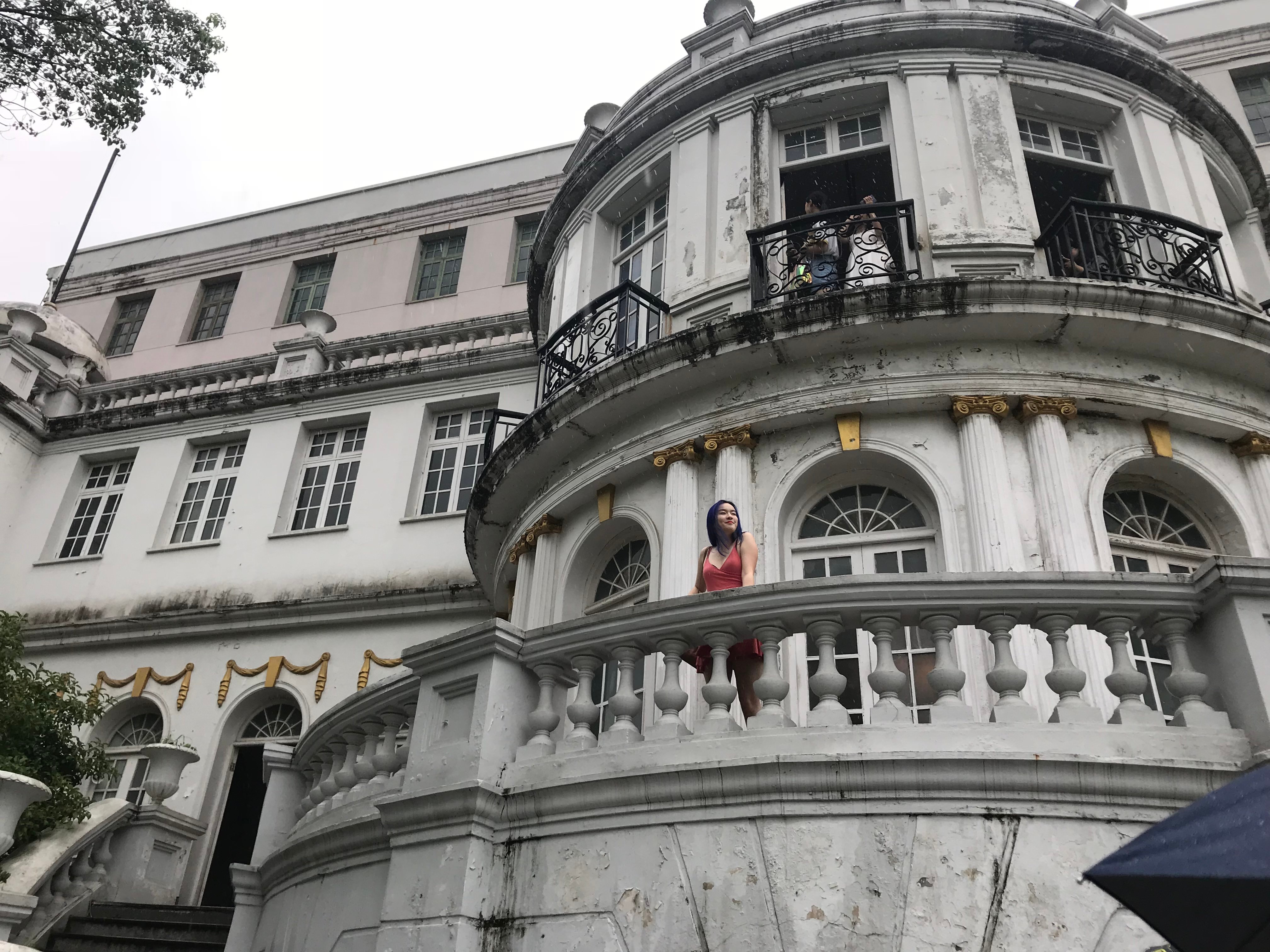 A woman in an orange slip dress gazing out from a second-floor stone balcony 