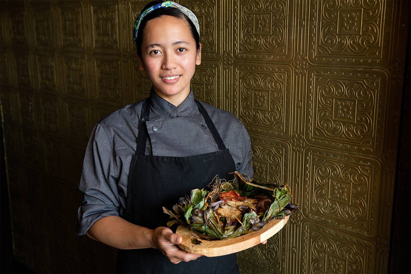 A smiling person of Filipino descent wearing an apron and holding a wooden plate containing a fish dish