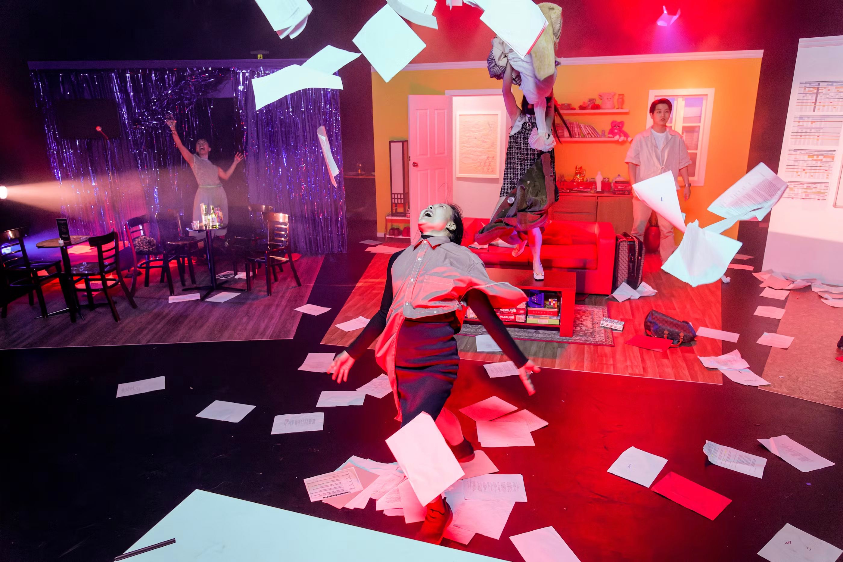 Actors perform on a vibrant stage set resembling a living room and café, with dramatic lighting and papers flying through the air in a chaotic, expressive scene.