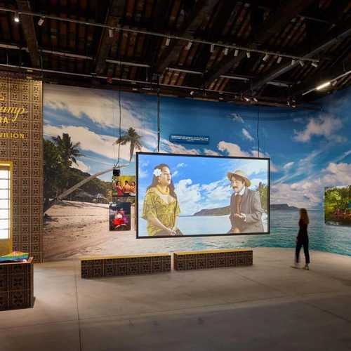 A colourful exhibition space with a full-wall mural of a beach in the Pacific and a video work projected on a screen.