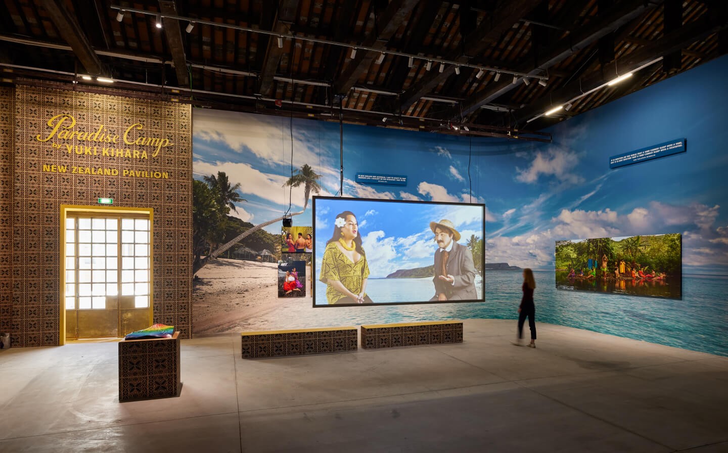 A colourful exhibition space with a full-wall mural of a beach in the Pacific and a video work projected on a screen.