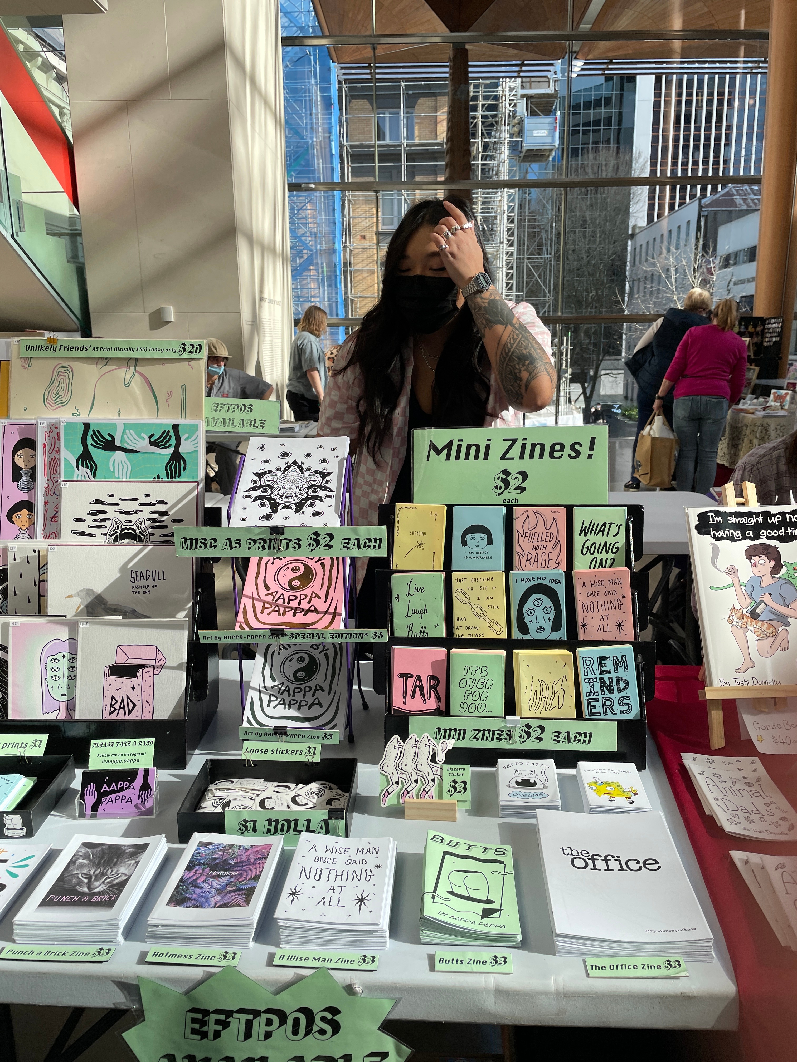 Photo of a stall of zines with a woman behind it wearing a face mask