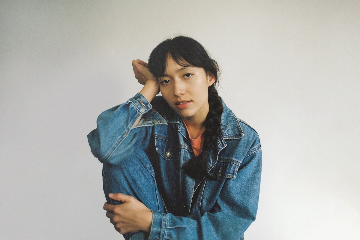 A woman gazing at the camera in a leather jacket and jeans, her hair in plaits