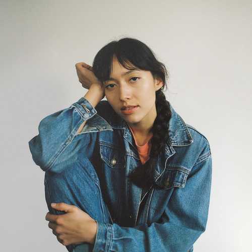 A woman gazing at the camera in a leather jacket and jeans, her hair in plaits