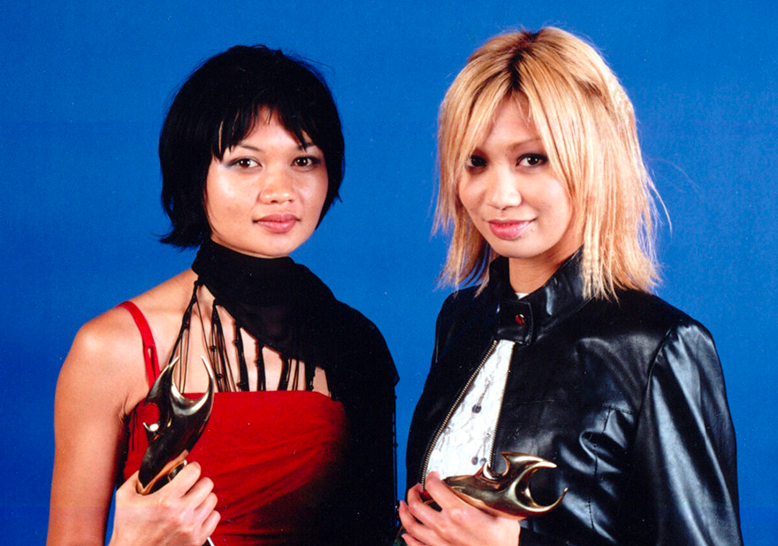 A woman with short black hair, a scarf and a red dress holding an award with a woman in a bleached shaggy bob and a leather jacket, also holding an award