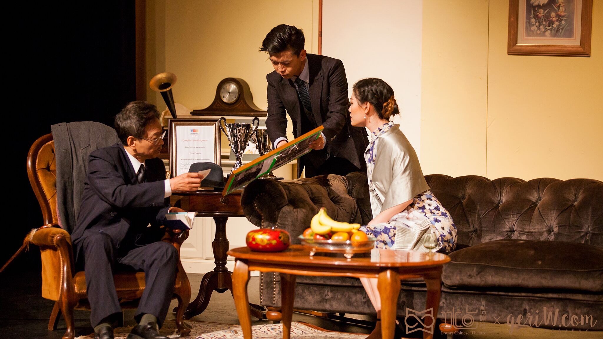 A lounge in which a man in a suit sits on a leather armchair while a couple show him a painting