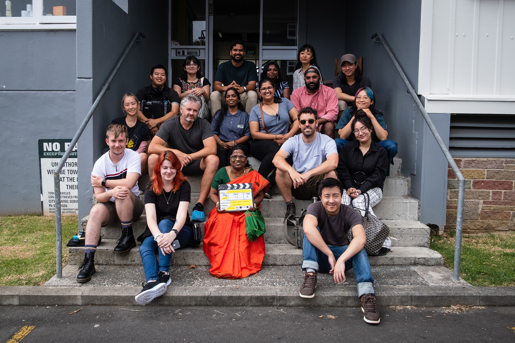 A group of 18 men and women sit outdoors on the entrance sets to a building. In the front centre, a women in a bright orange and green sari holds a film clapperboard.