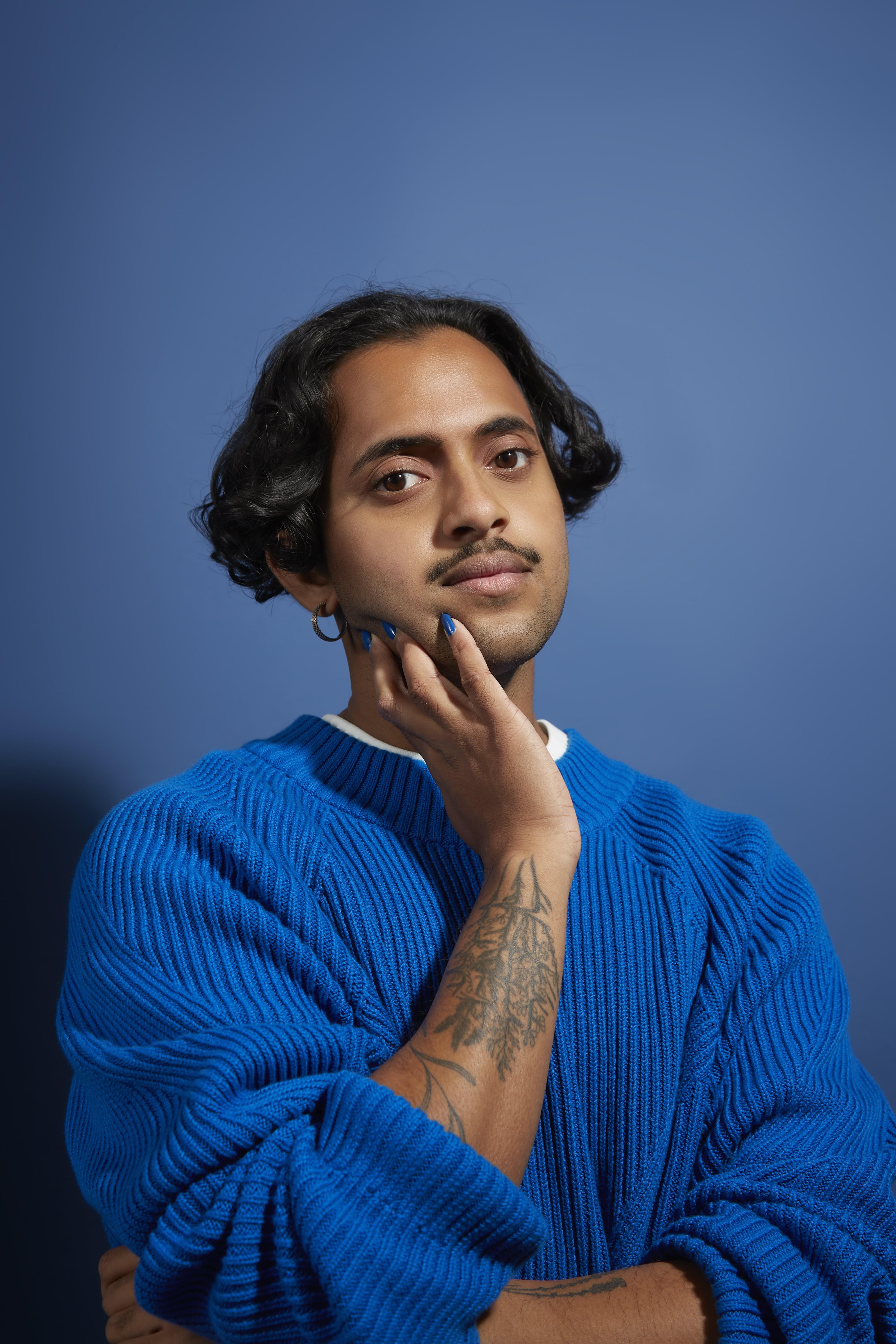 A man with cropped black hair and an earring, wearing a blue sweater, holds his face in his right hand.