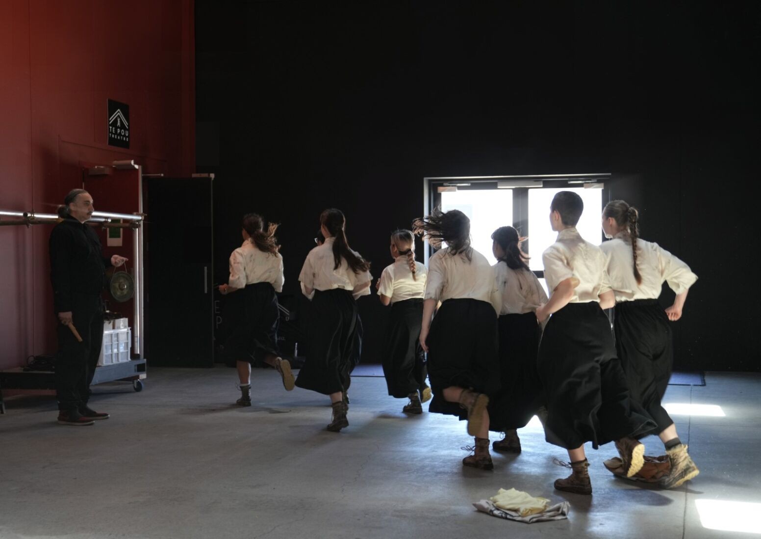 seven young girls wearing 1920s Taiwanese school uniform running towards the back, entering a theatre space. A man wearing police uniform staring at them on the side.