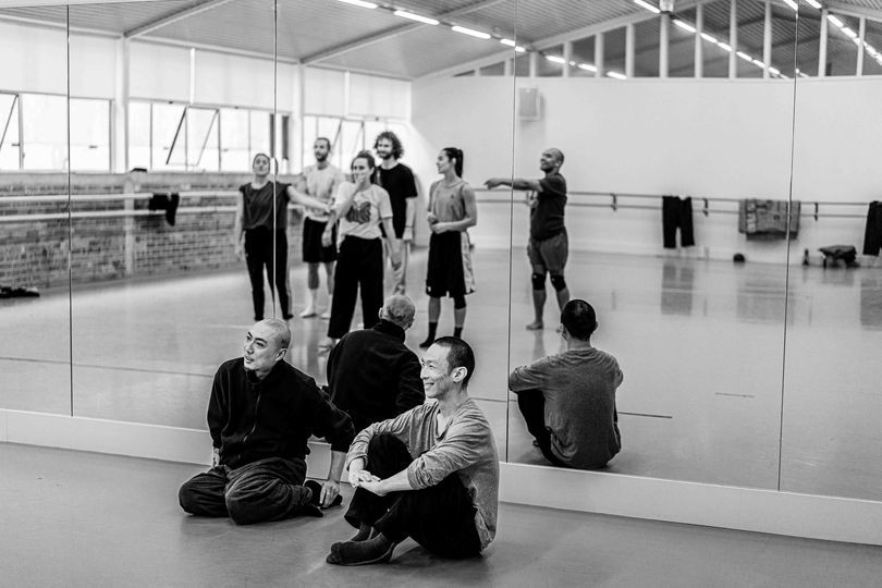 A group of dancers look into the mirror while two men sit cross legged facing them, smiling