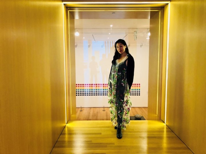 Yuk King Tan has long dark hair, worn loose, and wears a green patterned dress while posing at the end of an art gallery corridor.