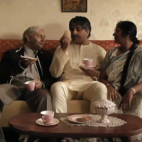 In a scene from ‘A Thousand Apologies’, two men and a woman sit on a sofa, drinking tea.