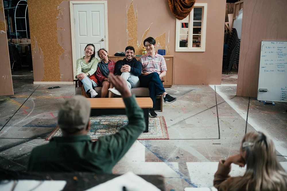 Four Asian young people sitting on a couch on a set, while others direct them.