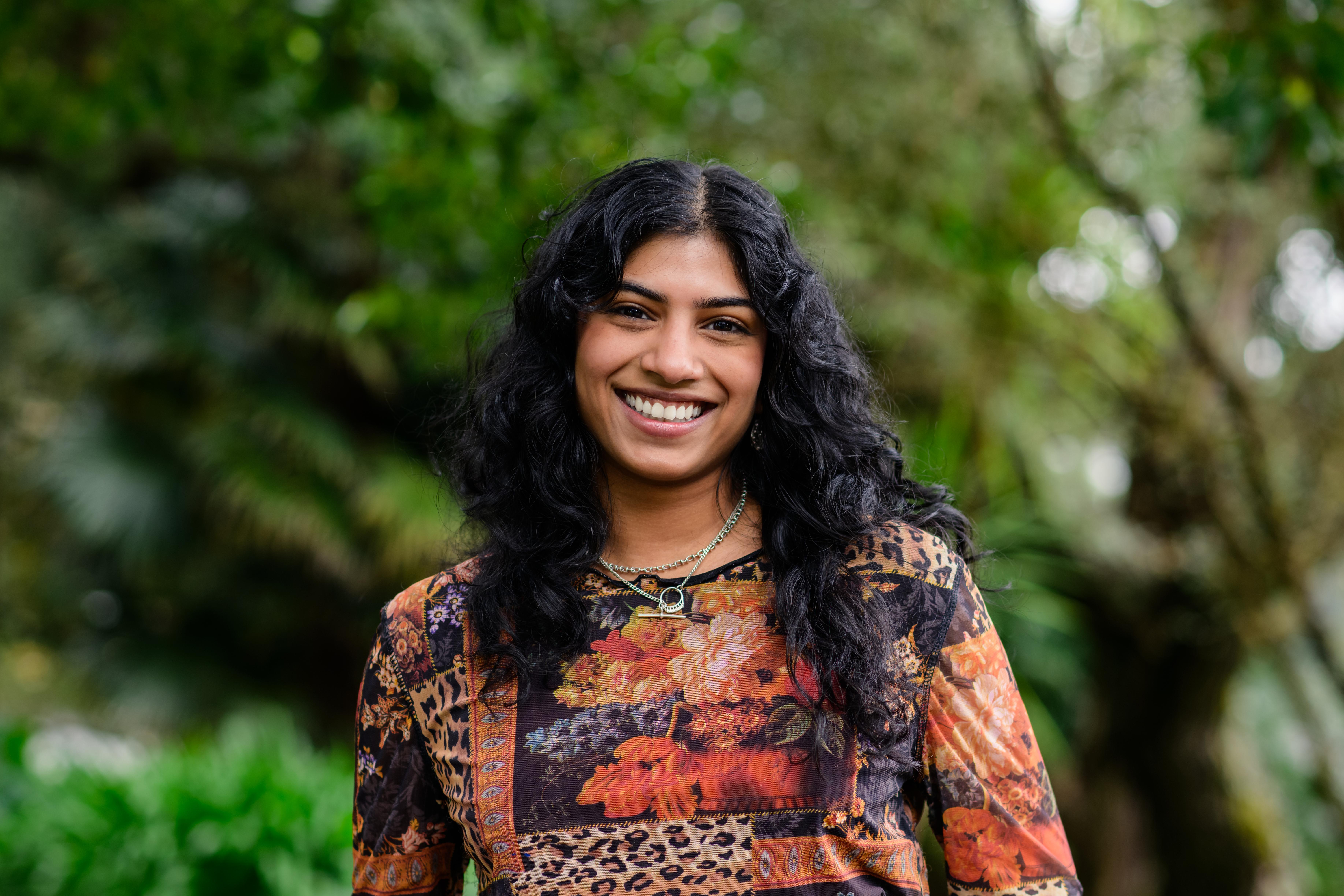 Dilohana has curly dark hair and is smiling widely, while posing outdoors for the photo.