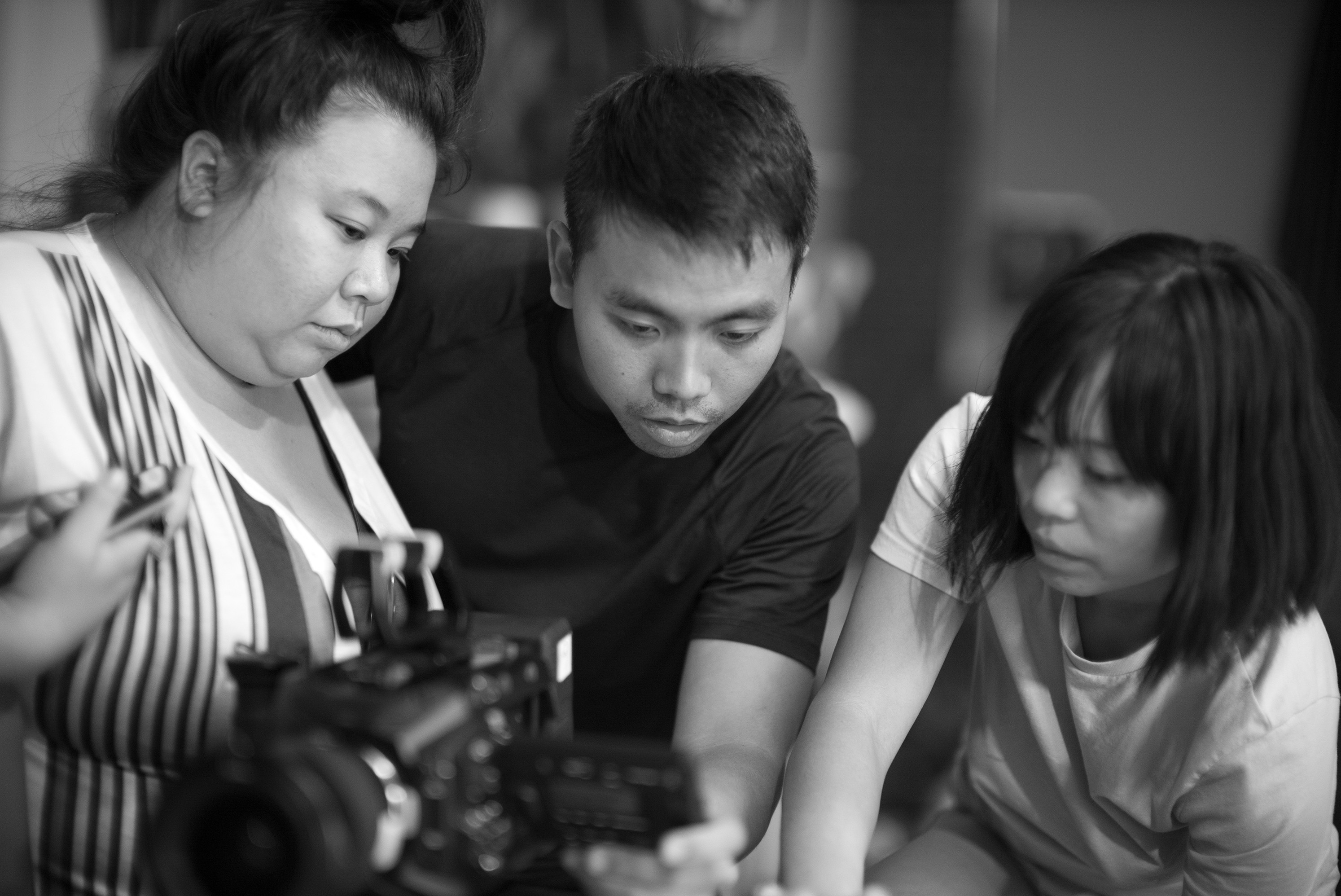 Black and white behind-the-scenes image of a film shoot, showing two women surrounding a man behind a digital video camera