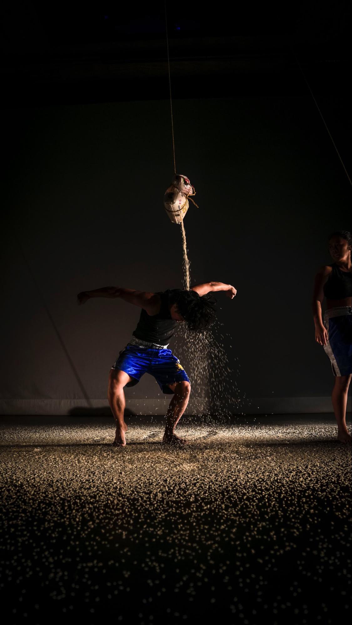 Action photo of a dancer with arms outstretched dancing underneath a container pouring grains of sand on their neck
