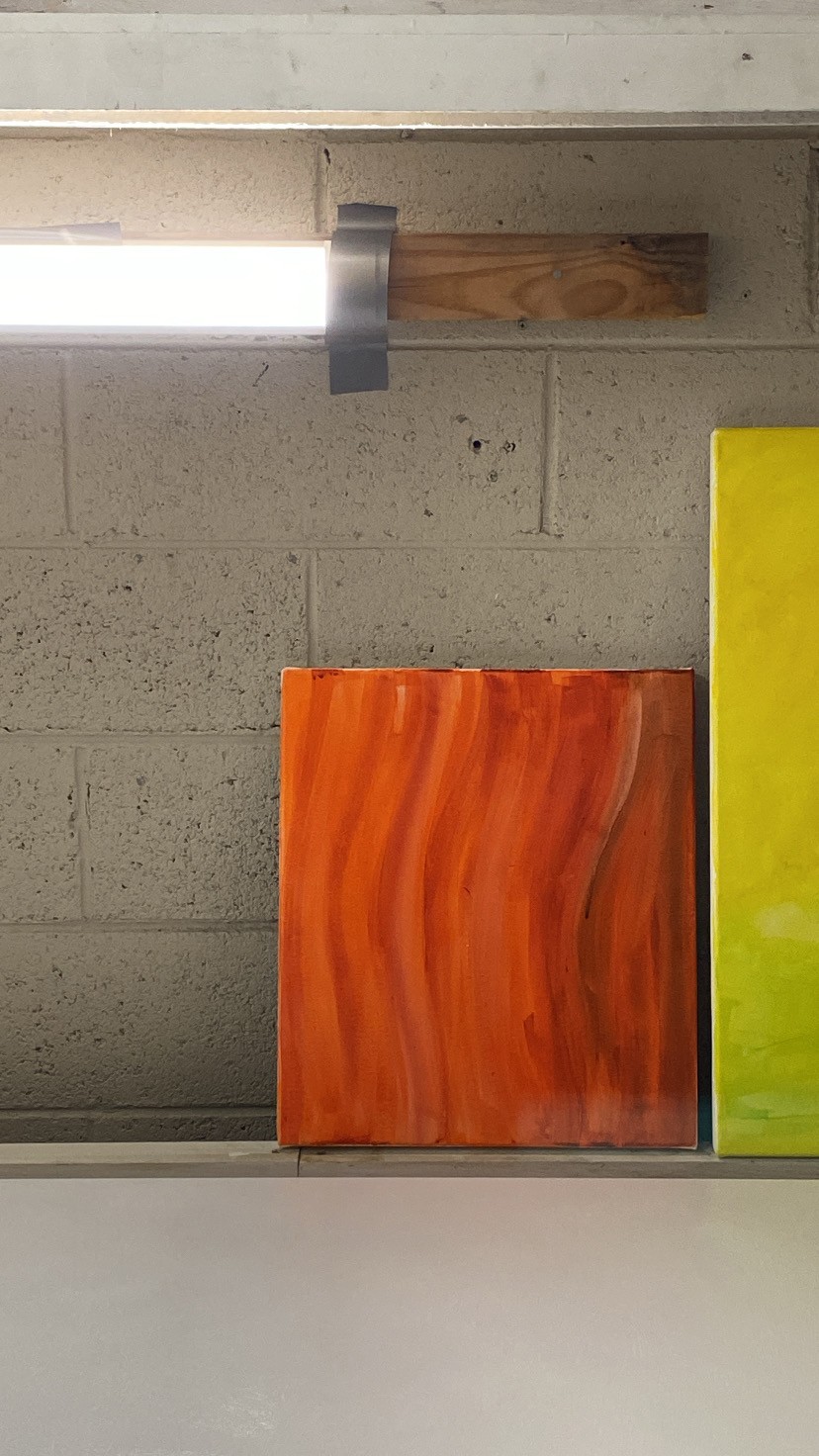 Small orange-red painting with undulating vertical brushstrokes sits on a ledge, with the edge of another slightly larger yellow and green painting next to it, cropped at the edge on the right side. The surface against which the paintings lean is dull white/grey brick, and there is part of a horizontal fluorescent light affixed to the surface with a piece of wood at the top left of the image is also cropped.