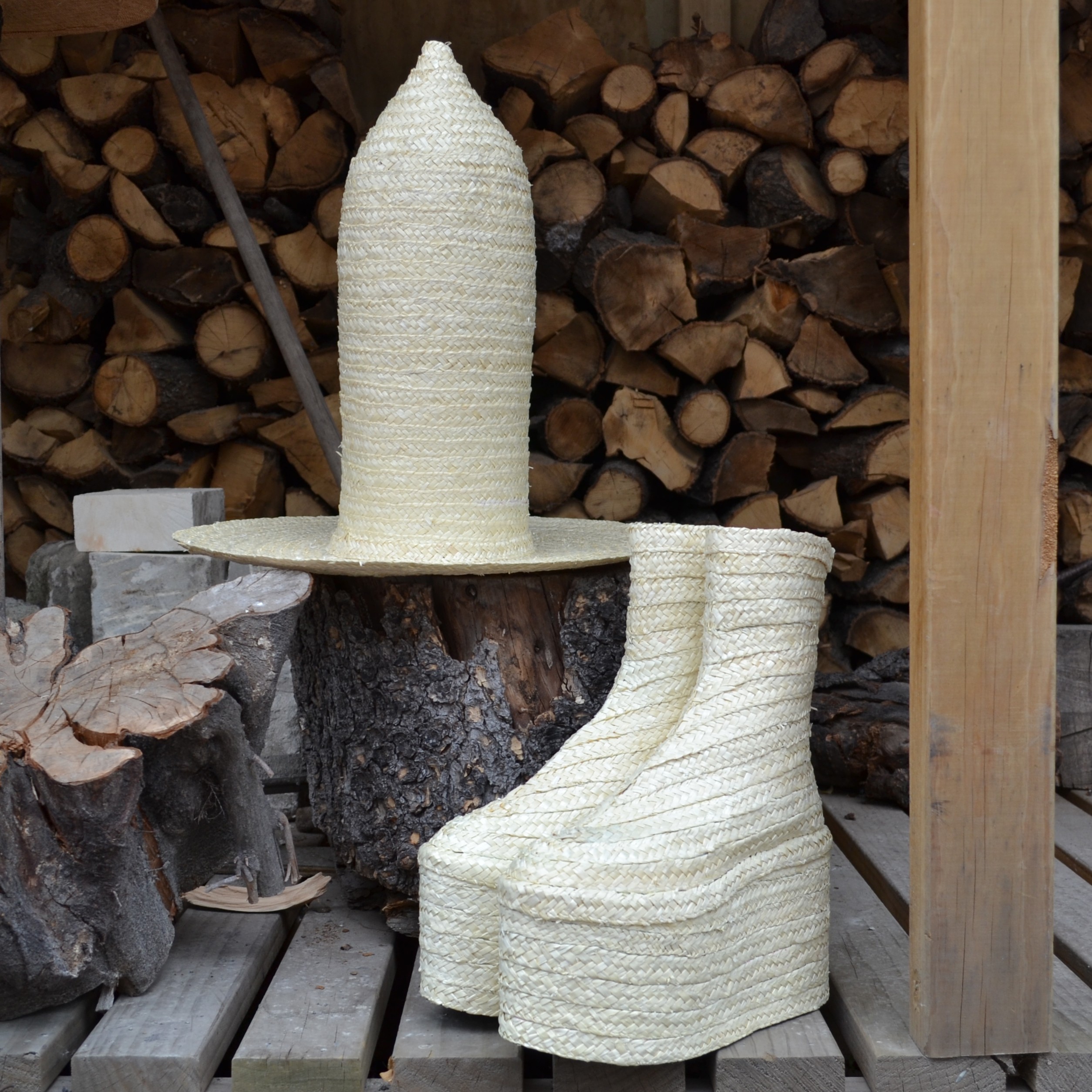 A tall hat and platform boots, both made of straw, sit amongst firewood