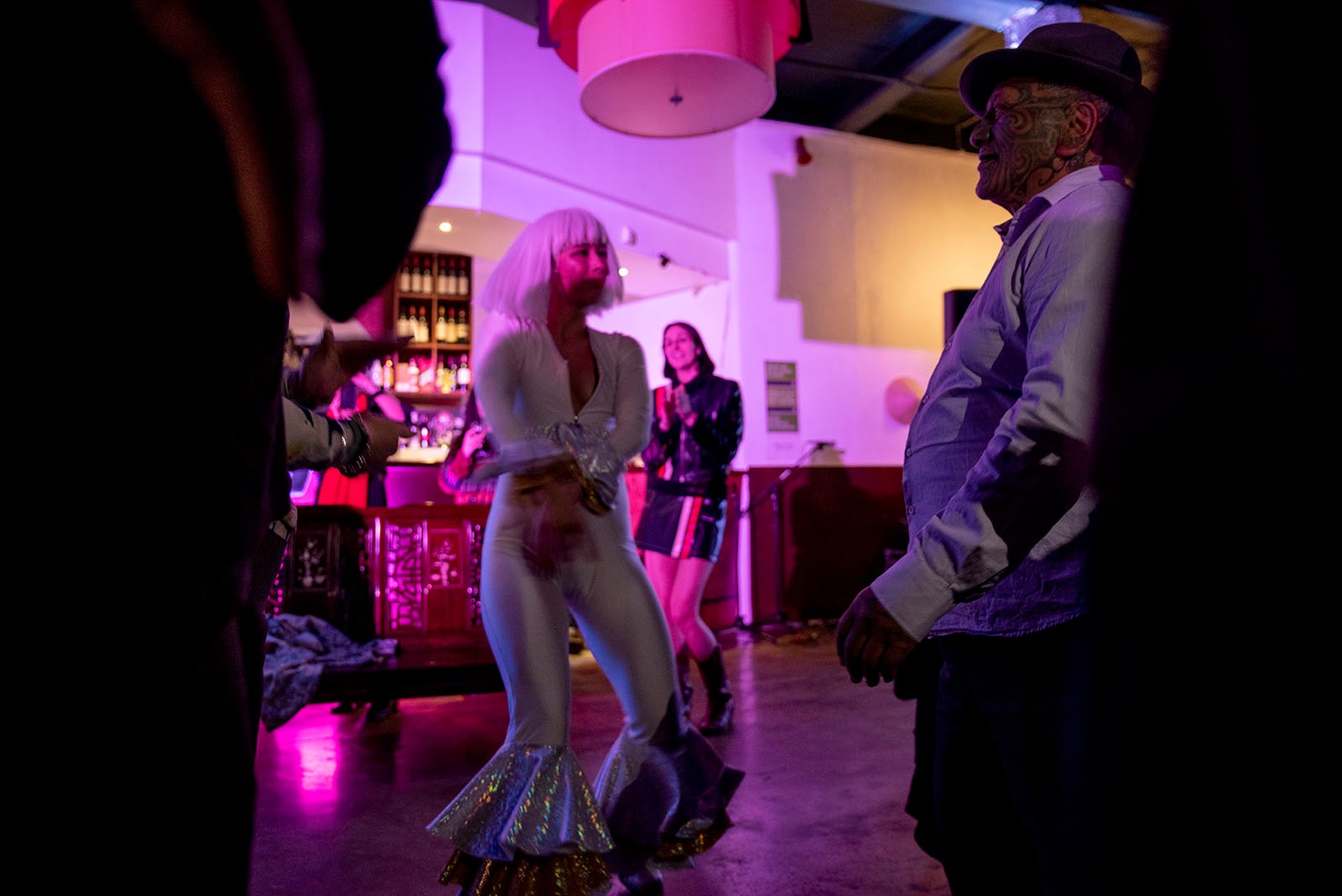 Emiko Sheehan dances in a white spandex suit with a white wig, with Tame Iti, inside a pink-lit restaurant