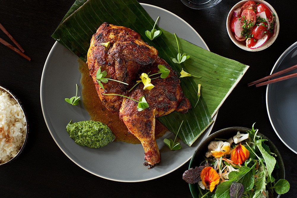 An artful flat-lay style photograph of well seasoned chicken on banana leaves in a grey plate surrounded by plates of salad and rice