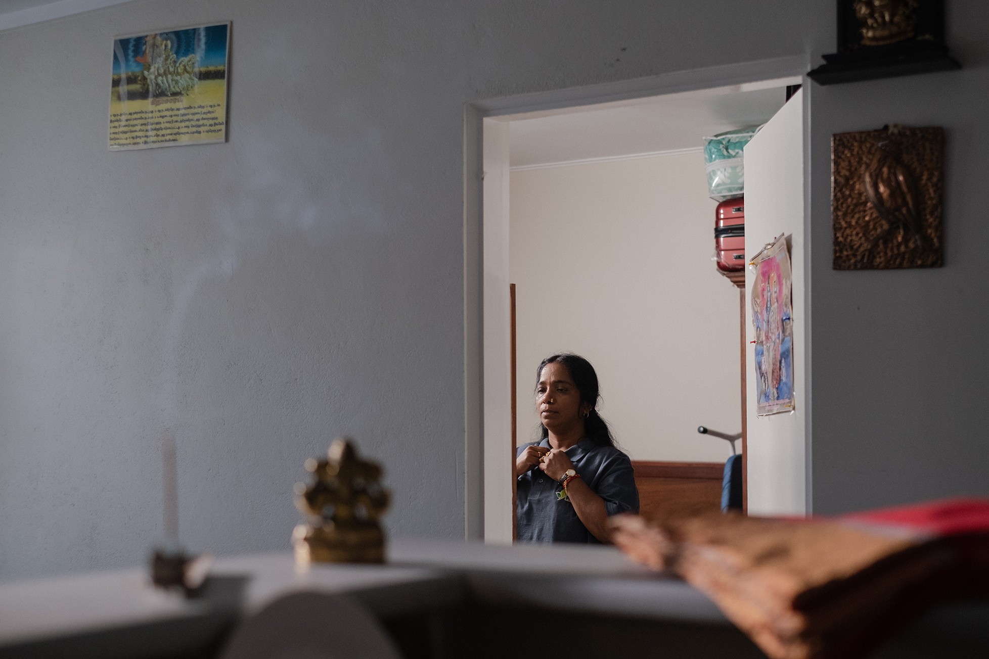 A woman stands in a doorway adjusting her shirt, with a soft focus on incense smoke and a small golden figurine in the foreground, within a sparsely decorated room.