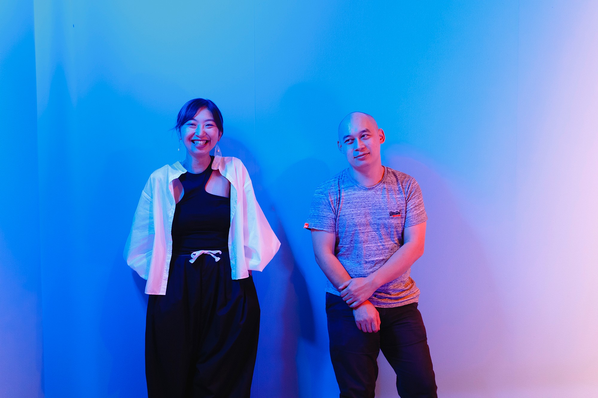 A woman in a black dress and a man in an grey shirt stand against a blue and pink lit wall