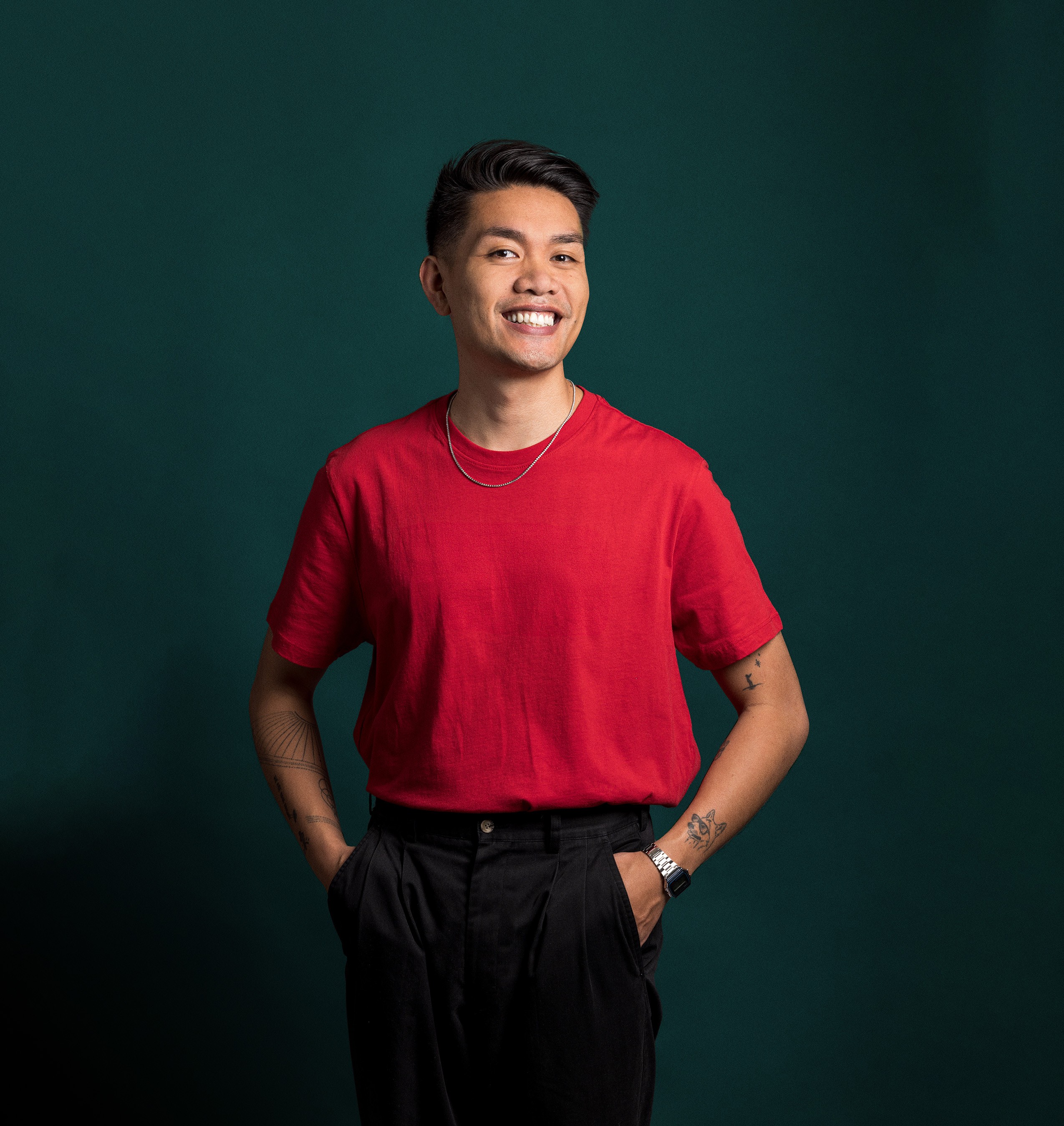 James Roque photographed smiling against a dark background, wearing a red t-shirt with his hands in his pockets.