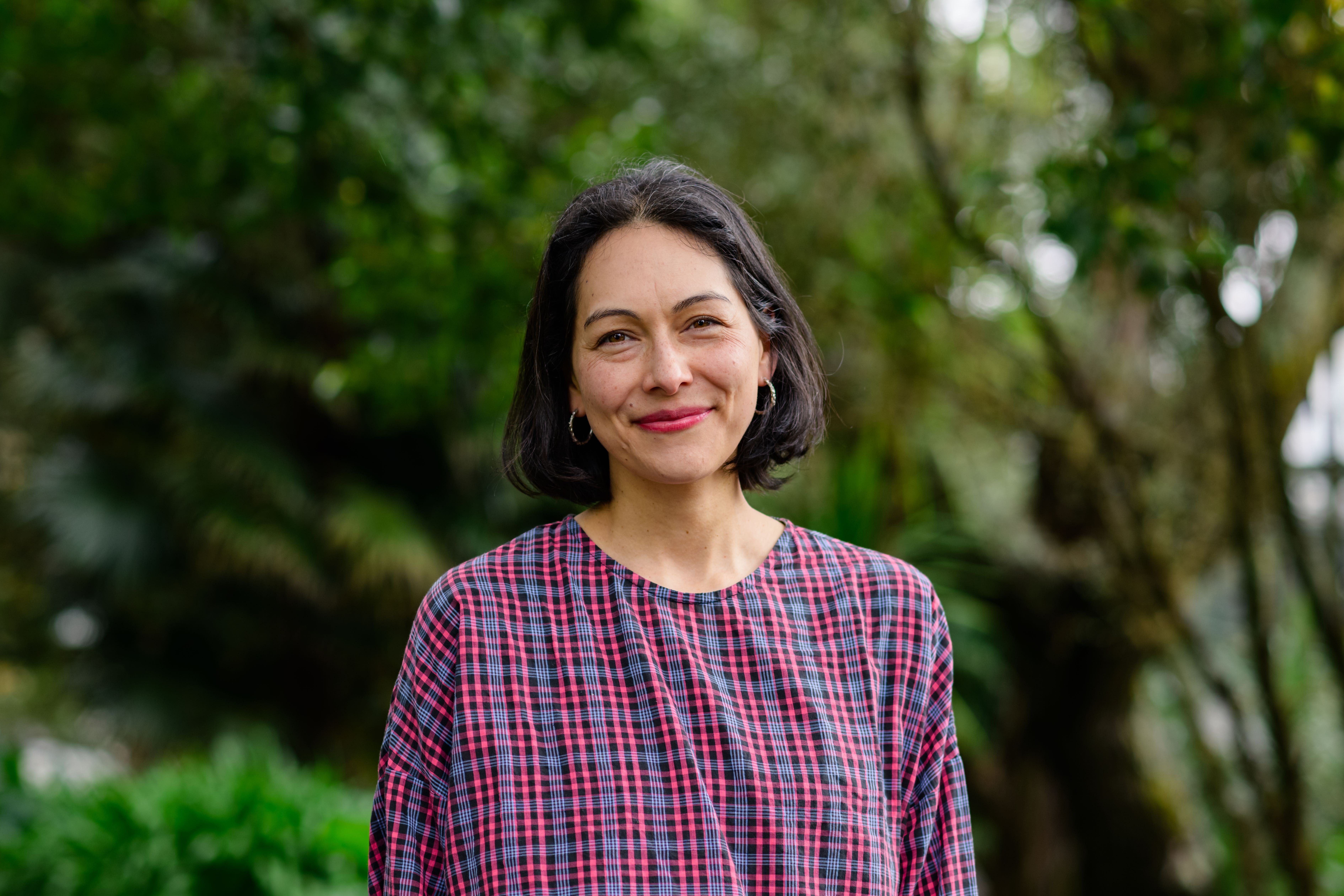 Melanie has a dark bob and is wearing small hoop earrings and checked shirt.Emma has long dark hair and is wearing a checked blue and brown dress. She poses outdoors for the photo.