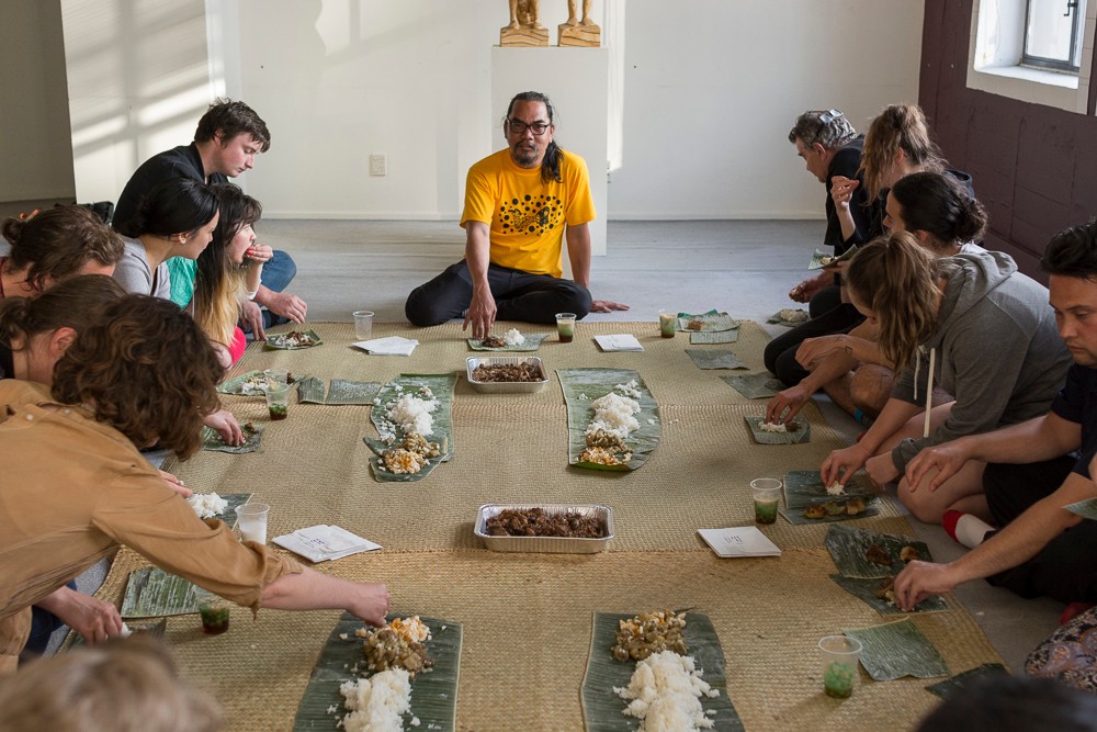 The Filipino artist wearing a yellow shirt sitting in the middle edge of a woven mat where various food has been placed. Surrounding the artist are other people who are sitting down and partaking of the food.